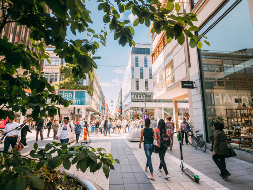 En grupp människor som går längs en gata bredvid butiker och höga byggnader i centrum.