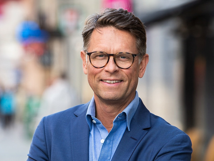 En man i blå kostym och glasögon står på en gata i en stad.